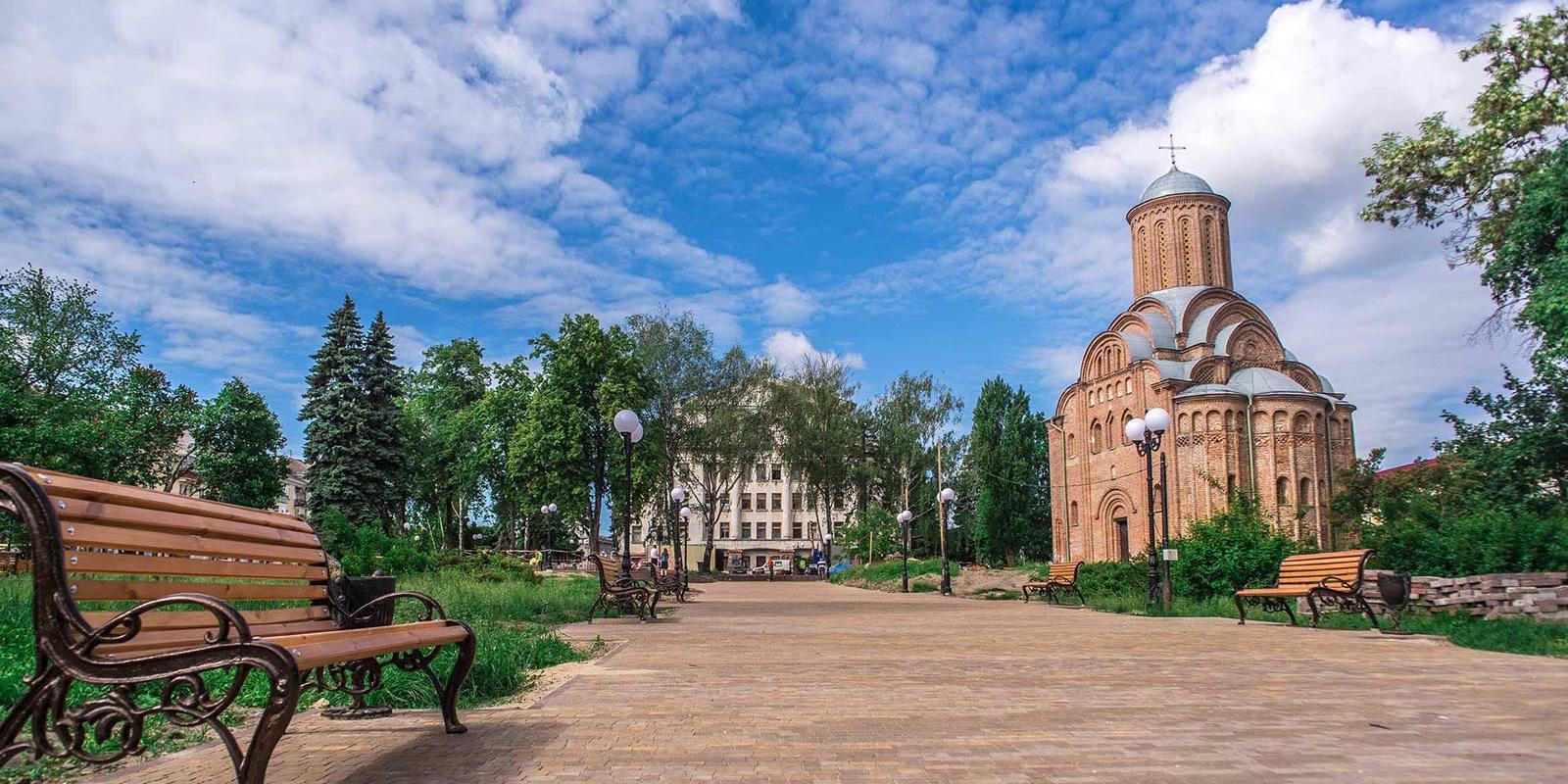 Парк з лавочками вздовж доріжок і церквою, а навколо зелені дерева та кущі